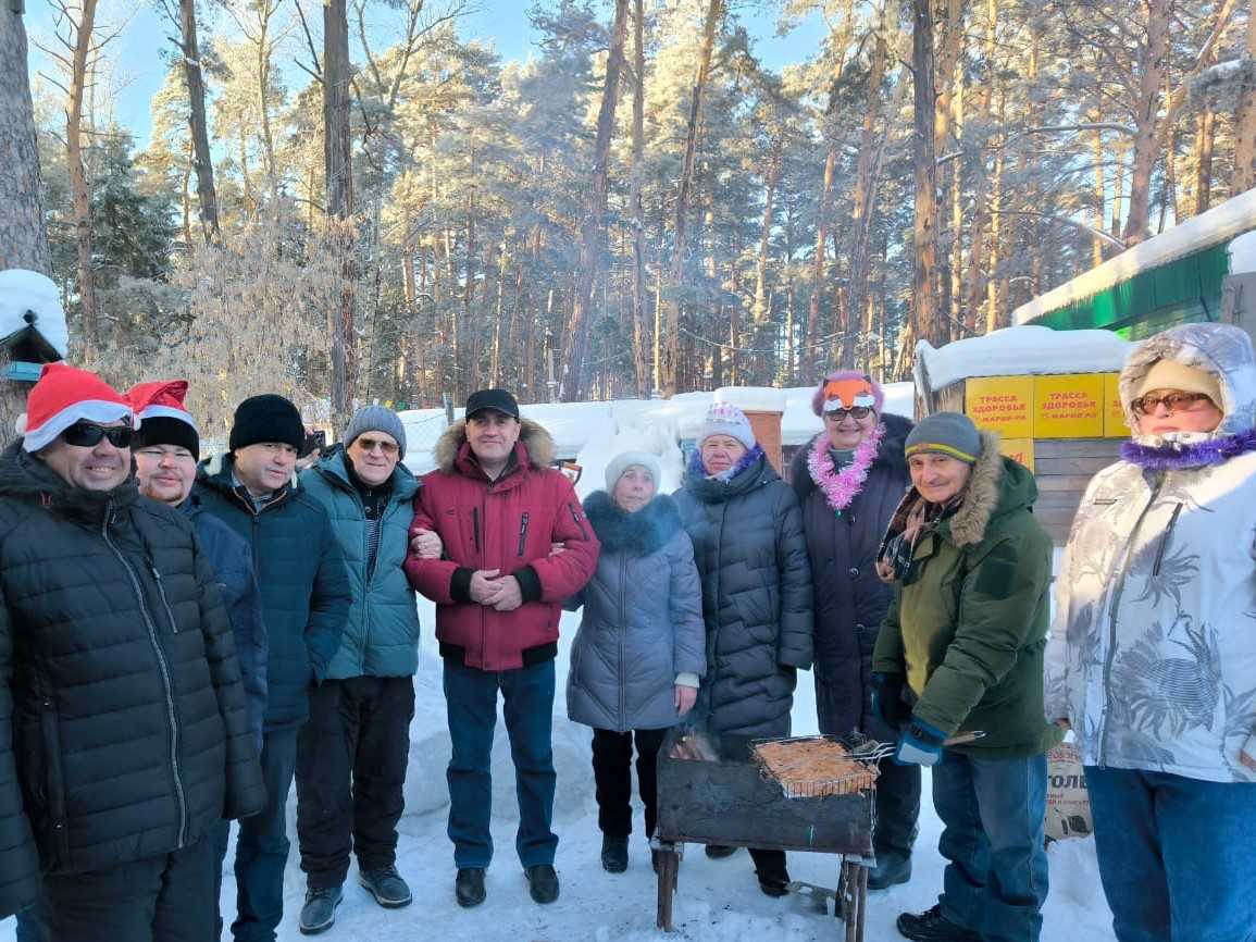 На фотографии изображена группа людей, стоящих полукругом на улице в зимней одежде. На переднем плане - мангал, с одной его стороны лежит несколько поленьев дров, а с другой стороны, на металлической решетке готовится мясо. Люди одеты в тёплые куртки и шапки, некоторые из них в шапках Деда Мороза. На заднем плане виден заснеженный лес, освещенный солнцем. Люди улыбаются и выглядят радостными. На заднем плане также виден баннер, но текст на нём неразборчив.